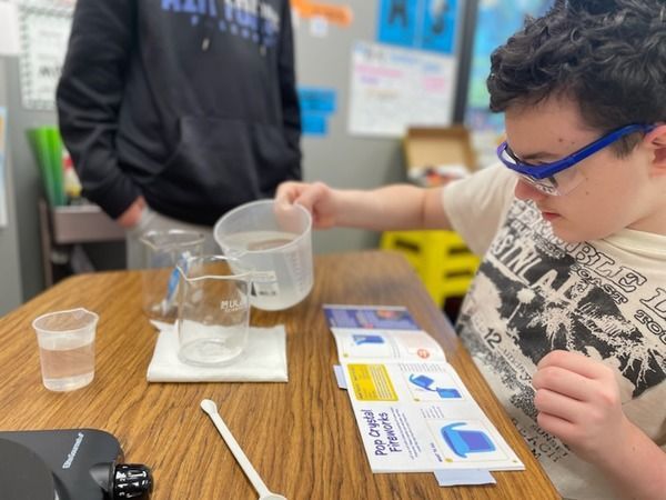 A boy wearing safety glasses is pouring water into a measuring cup.