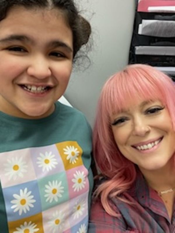 A woman with pink hair is smiling next to a girl wearing a shirt with daisies on it