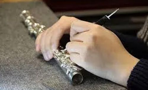 A person's hands are using a tool to adjust a silver flute on a gray surface.