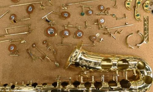 Disassembled saxophone with golden body, keys, pads, and mechanisms on a tan surface.