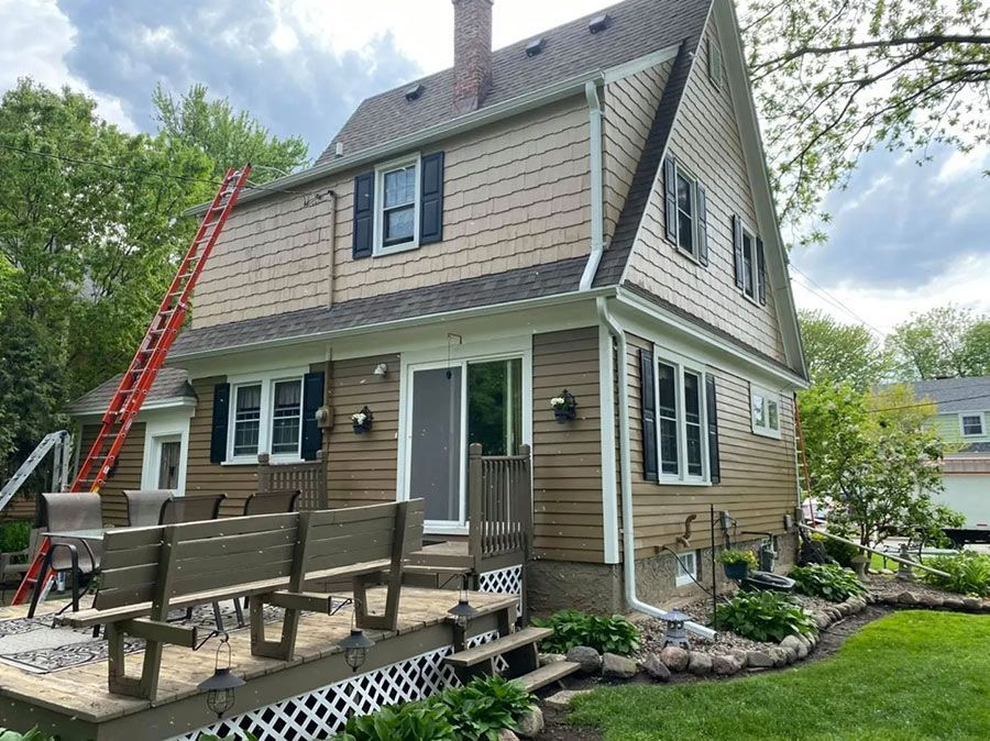 Tan house with a wooden deck and a ladder leaning against it, surrounded by trees and a green lawn.