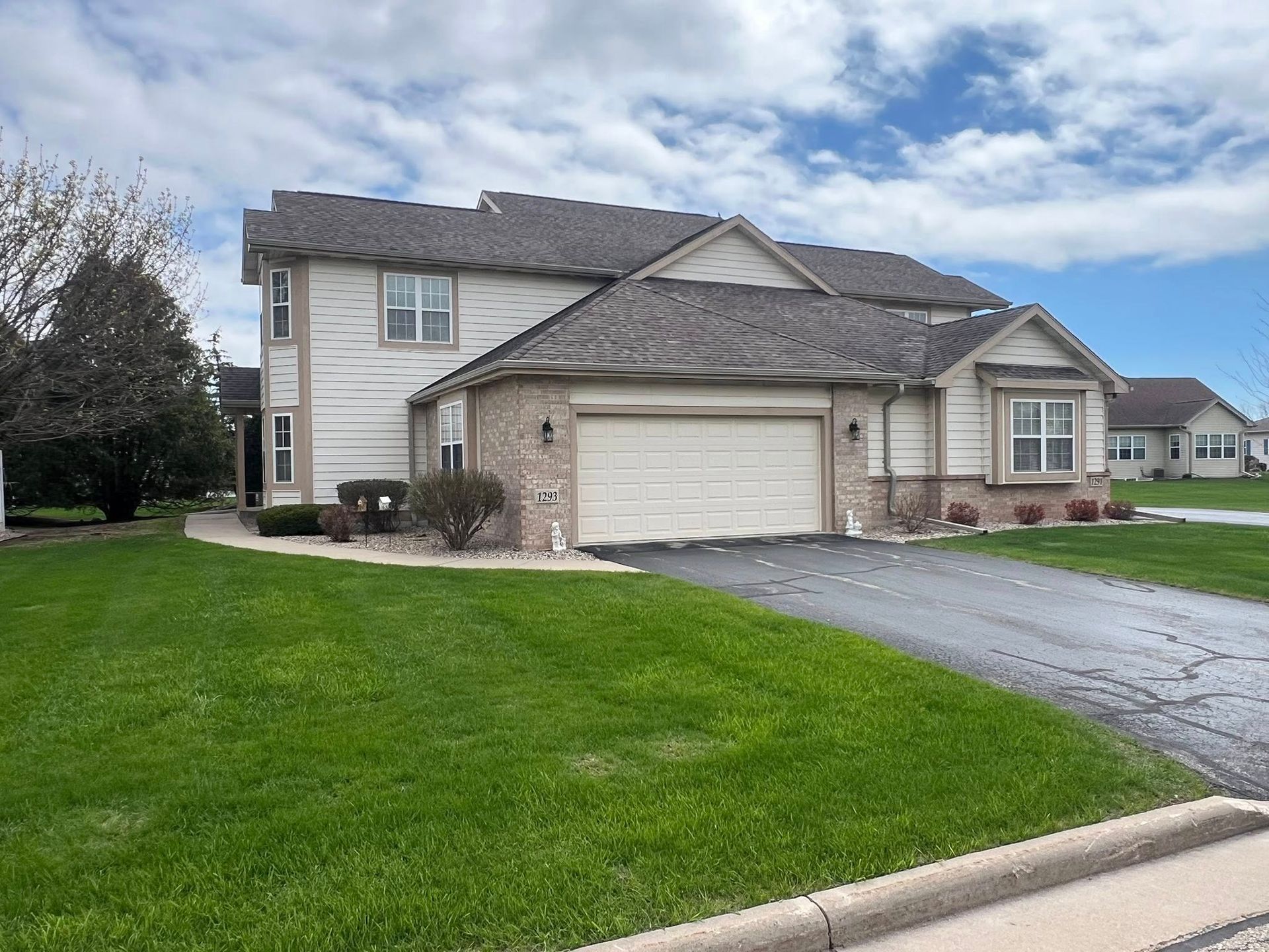 Two-story beige house with a brick facade and attached garage, situated on a green lawn with a black driveway.