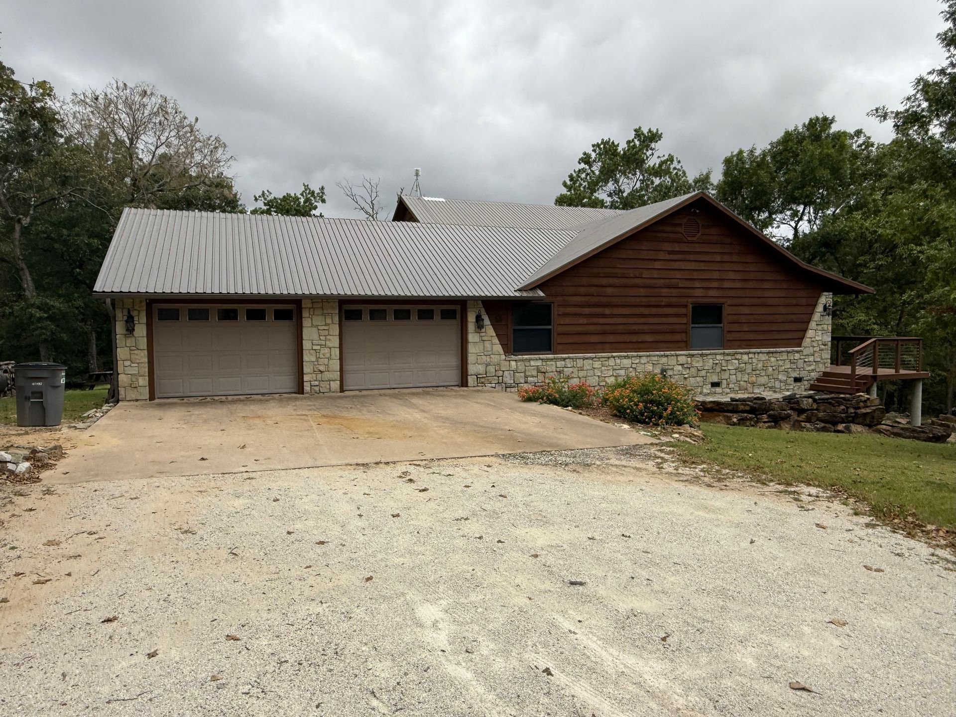 House with a gray metal roof, stone and wood siding, and two-car garage. Overcast sky.