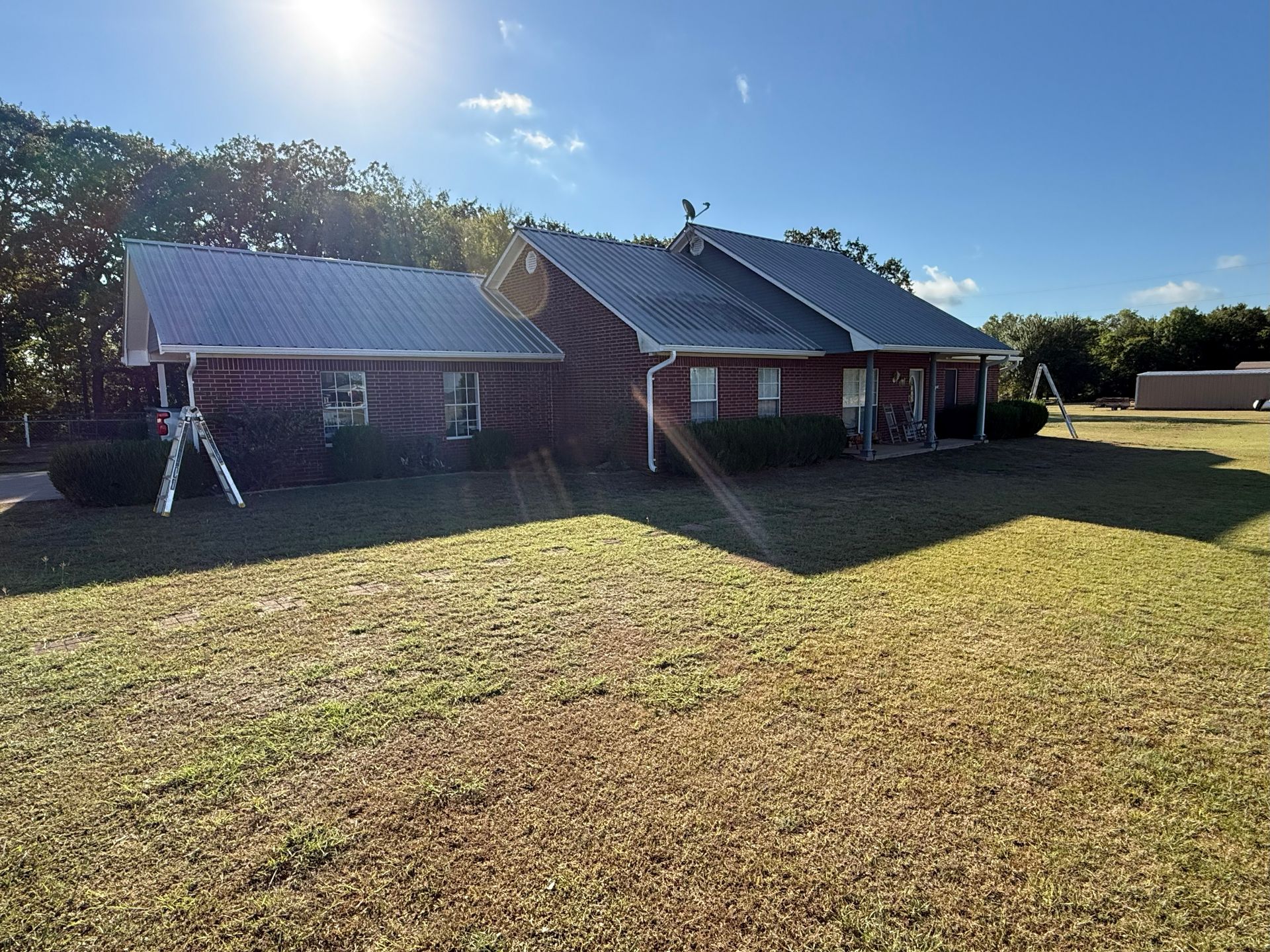 Brick house with a metal roof on a sunny day. Green grass and trees surround it.
