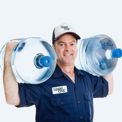 A man wearing a water one shirt is carrying two bottles of water