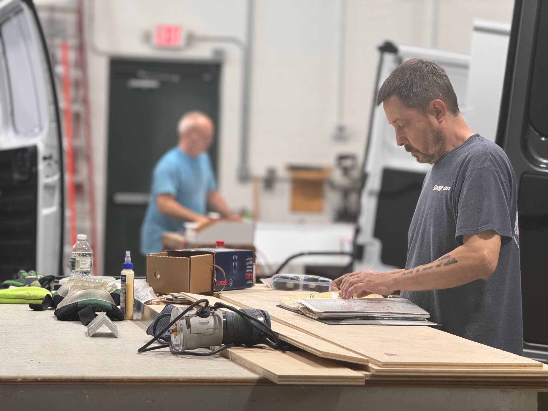 Worker measuring wood on a workbench in a workshop, with tools and another person blurred in the background.