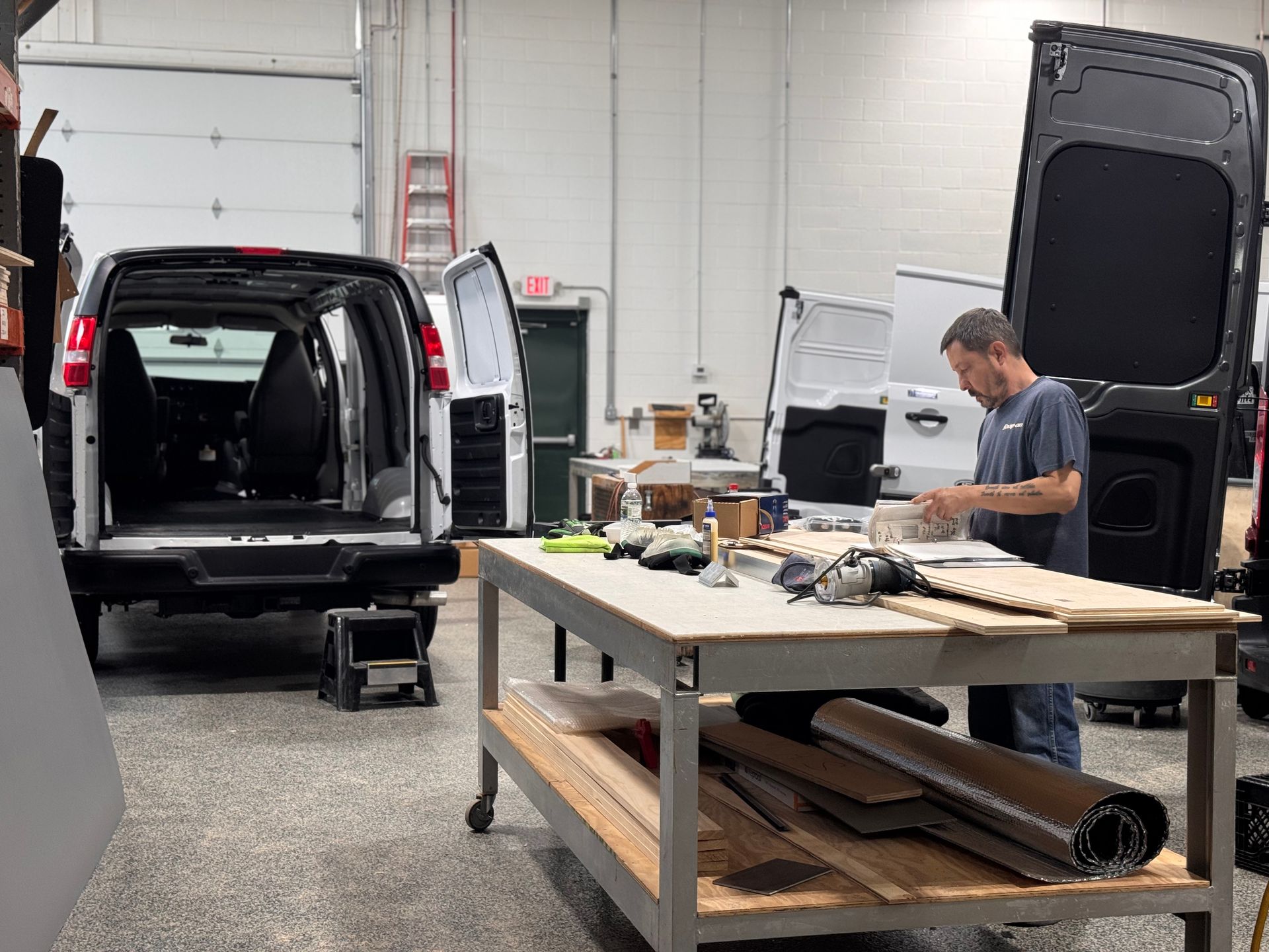 Man working on equipment in a garage workshop beside an open black van with tables and tools.