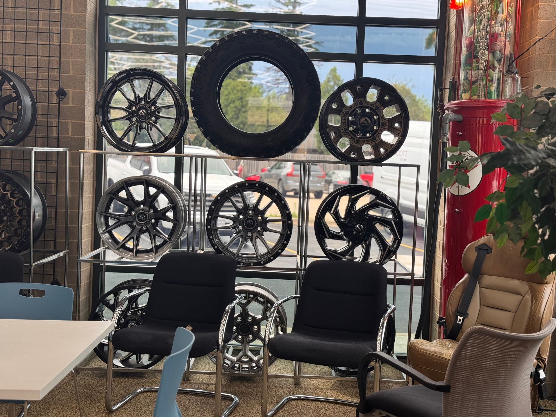 Tire display wall in a shop with black chairs, a table, and potted plant by a large window.