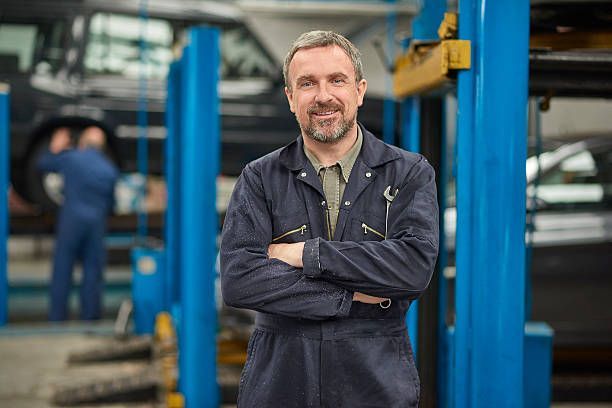 Mechanic with crossed arms in auto shop, smiling. Another mechanic works on a car in the background. Blue overalls.
