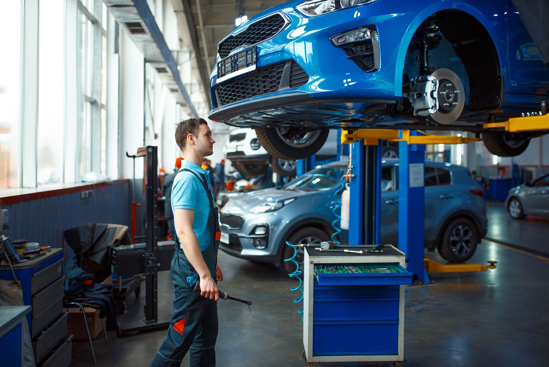 A man is standing in front of a blue car on a lift in a garage.