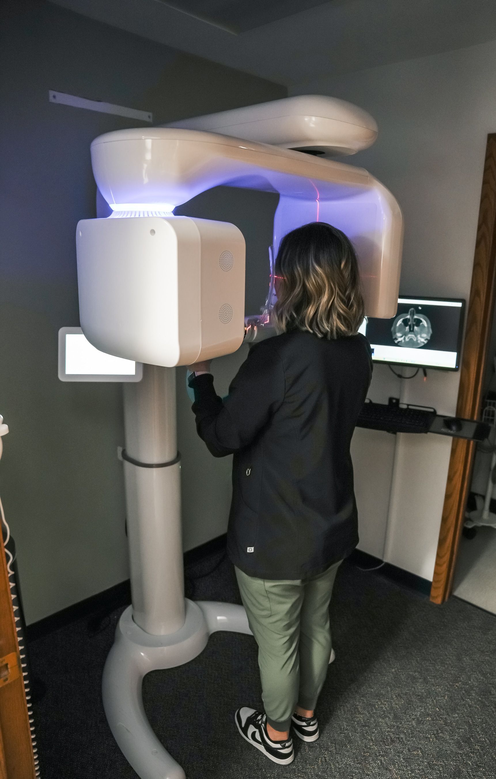 Woman operating dental X-ray machine in a clinic. She is wearing a black jacket and green pants.