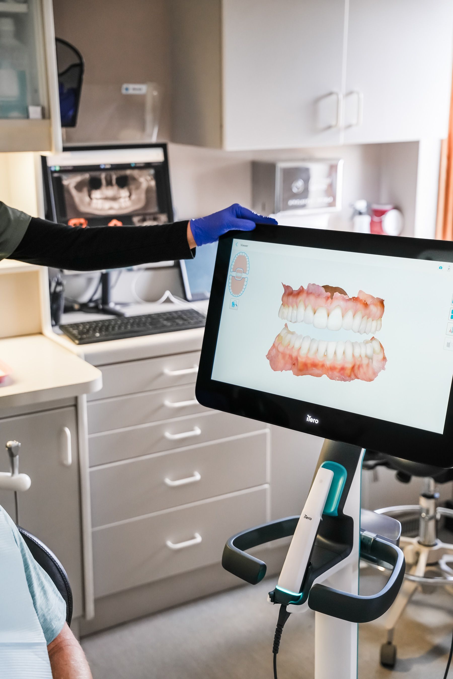 Dentist's office: Person points at 3D dental model on screen. X-ray of teeth visible on another screen.