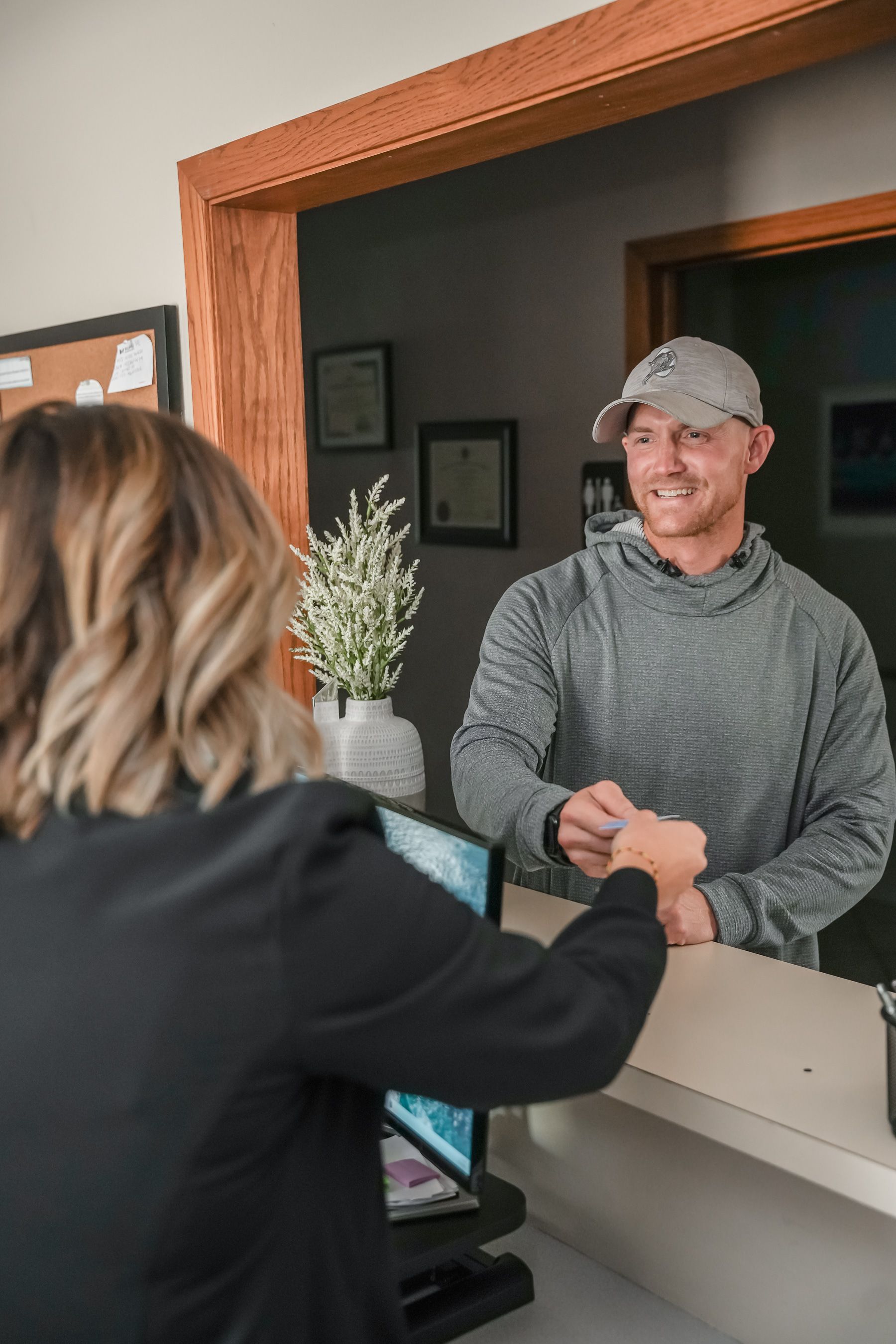 A woman at a desk is handing a card to a smiling man in a gray hoodie and baseball cap at a reception area.