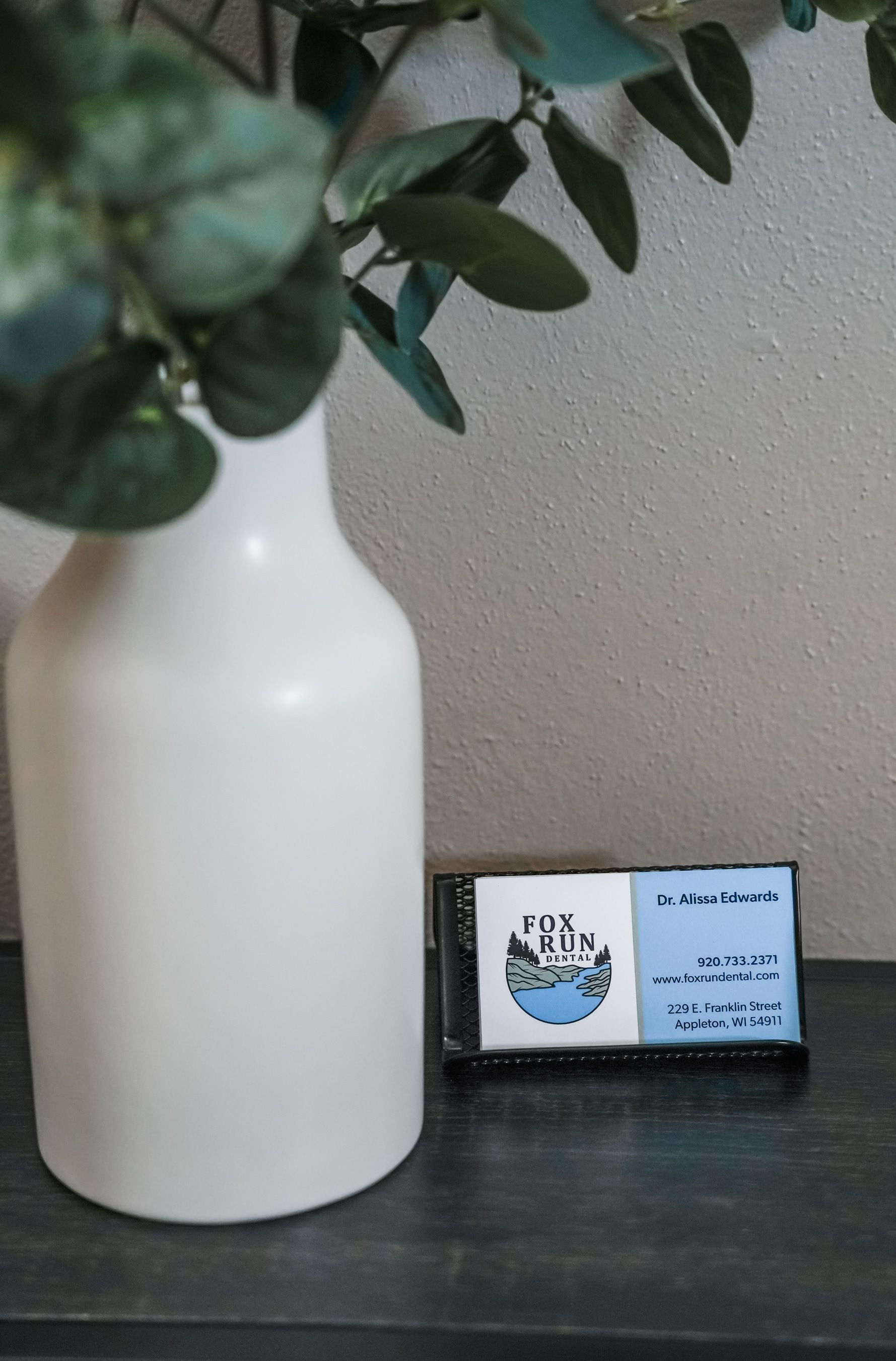 White vase with blue-green leaves, next to a soap bar with a logo on a dark blue surface.