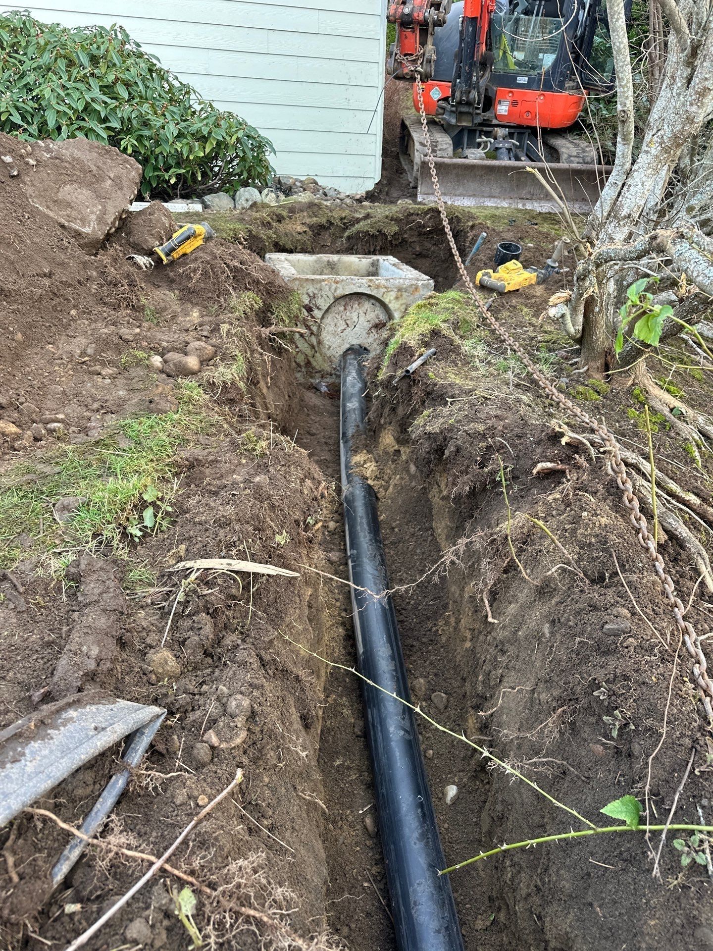 Trench with black pipe running through dirt toward a house, with an orange utility machine in the background.