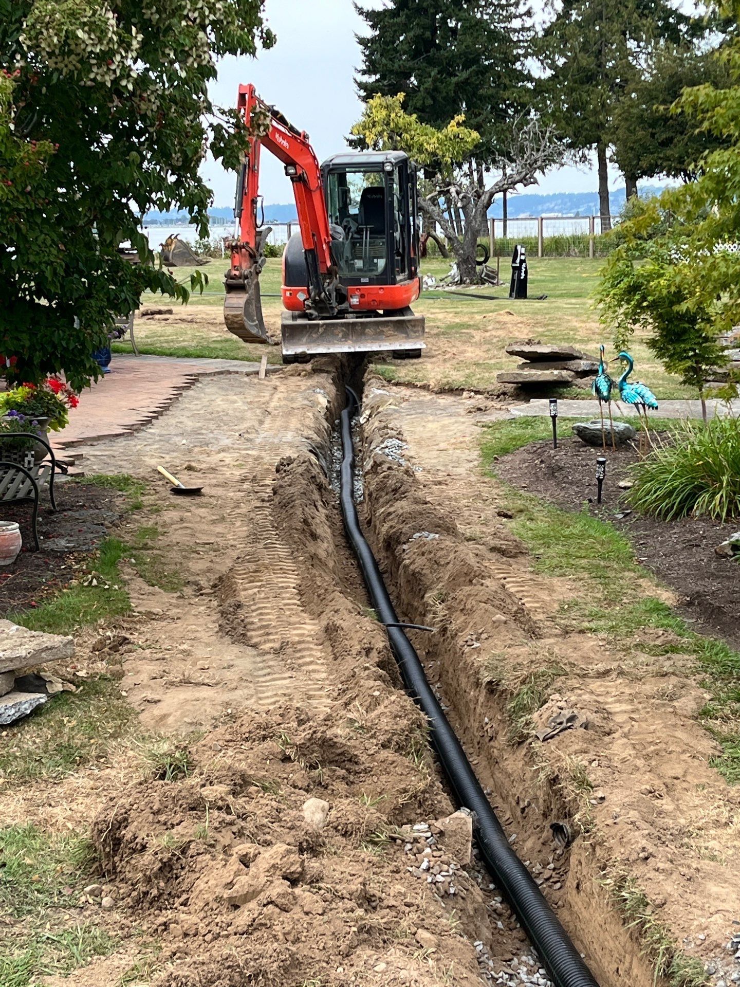 Excavator trenching a dirt path beside a black pipe in a park near trees and water.