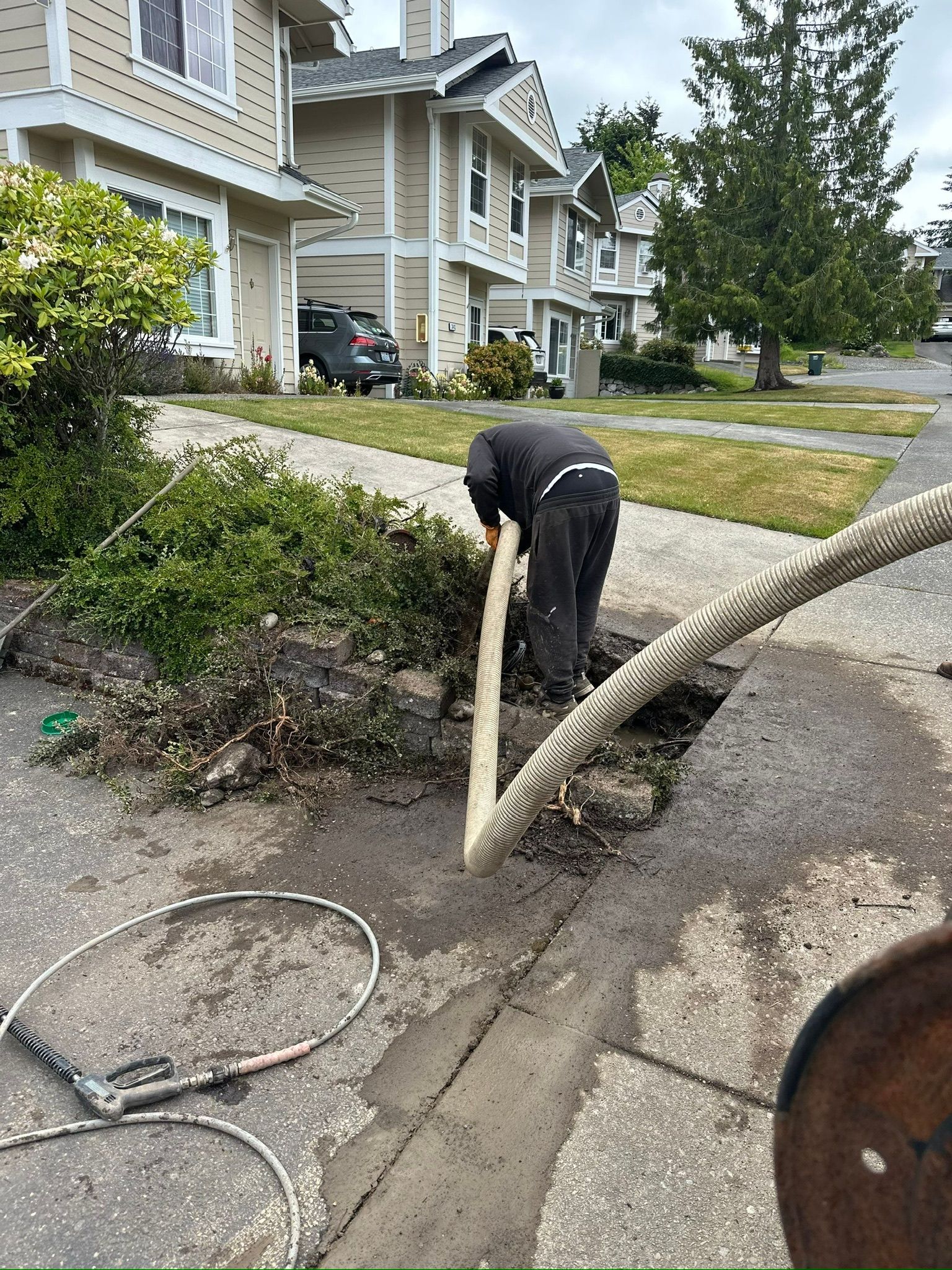 Worker using a hose to clean a curbside gutter in front of houses on a residential street.