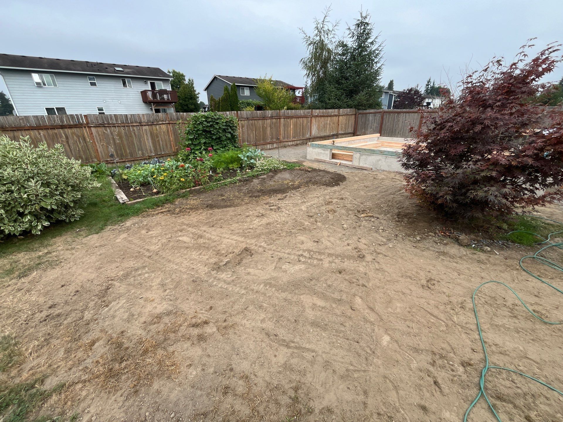 Bare dirt backyard with a wooden fence, raised bed, shrubs, and a garden hose under cloudy sky.