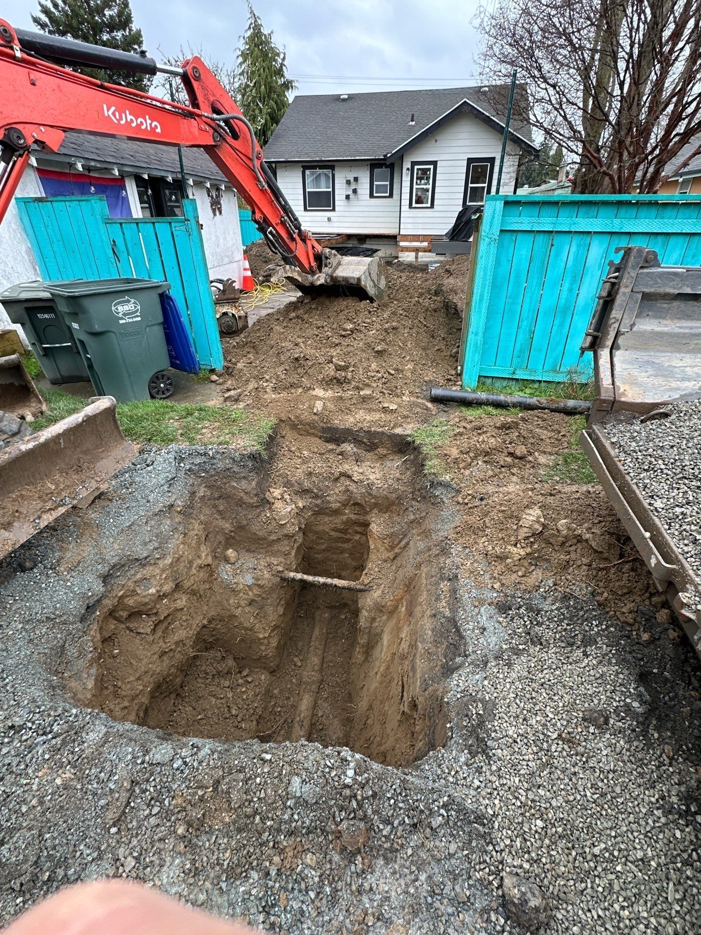 Backyard excavation with a deep trench, excavator, and blue fencing near a house.