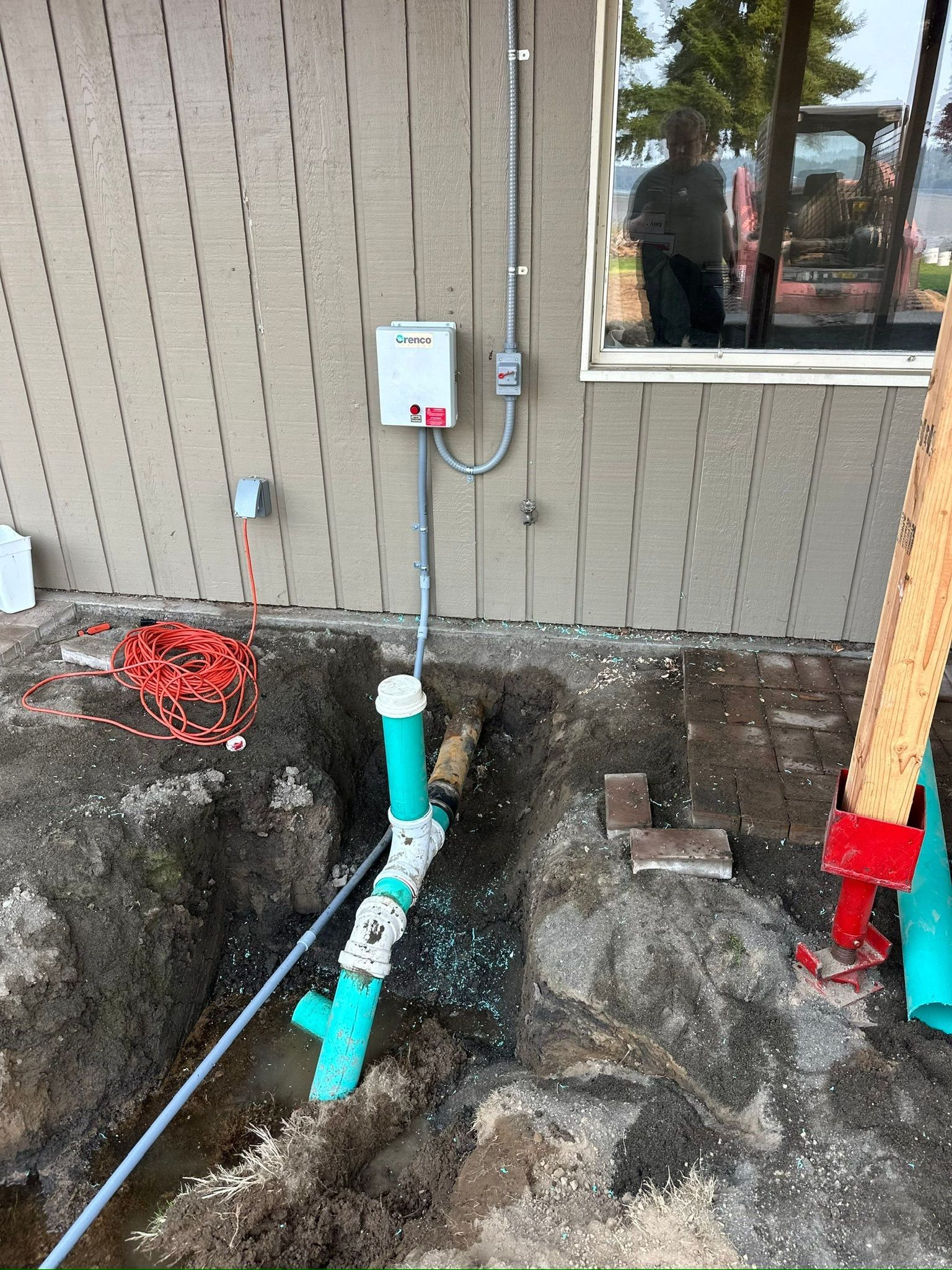 Outdoor house wall with exposed plumbing pipes and utility boxes near a dug-up trench.
