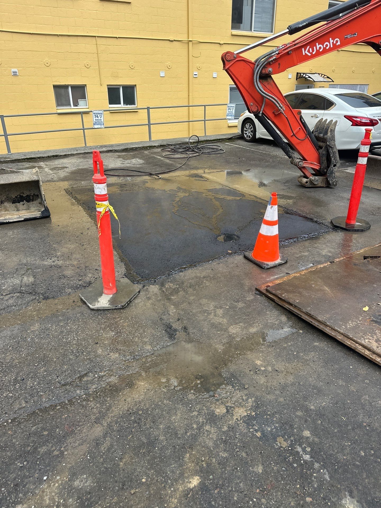 Construction site with orange cones around a patched road near an excavator and yellow building.