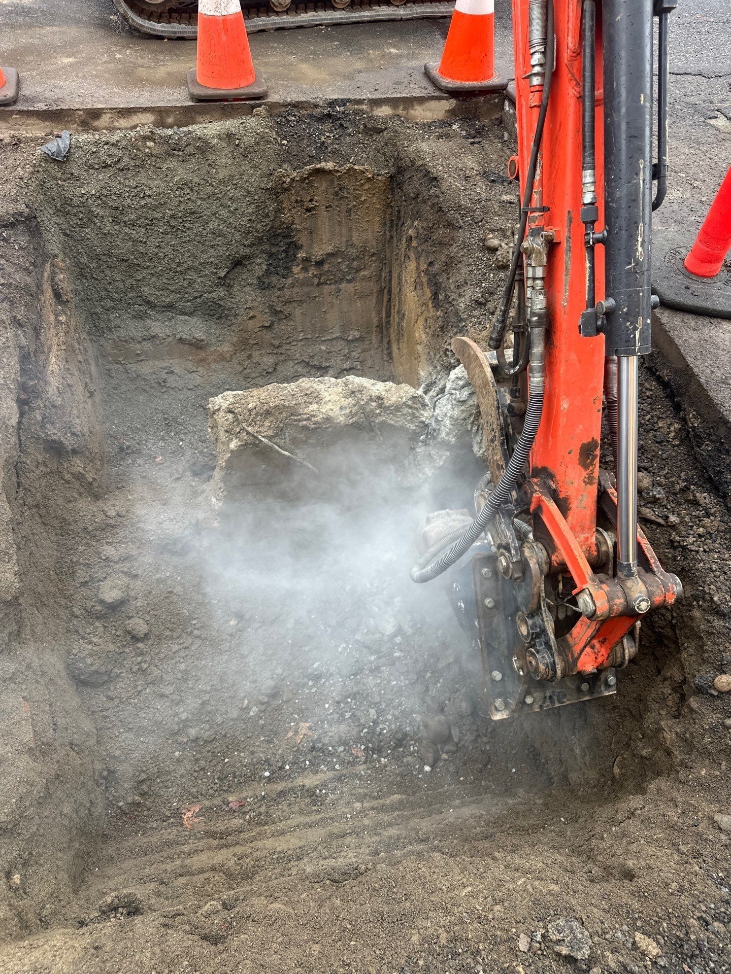 Excavator breaking concrete in a trench, with dust rising and orange safety cones nearby.
