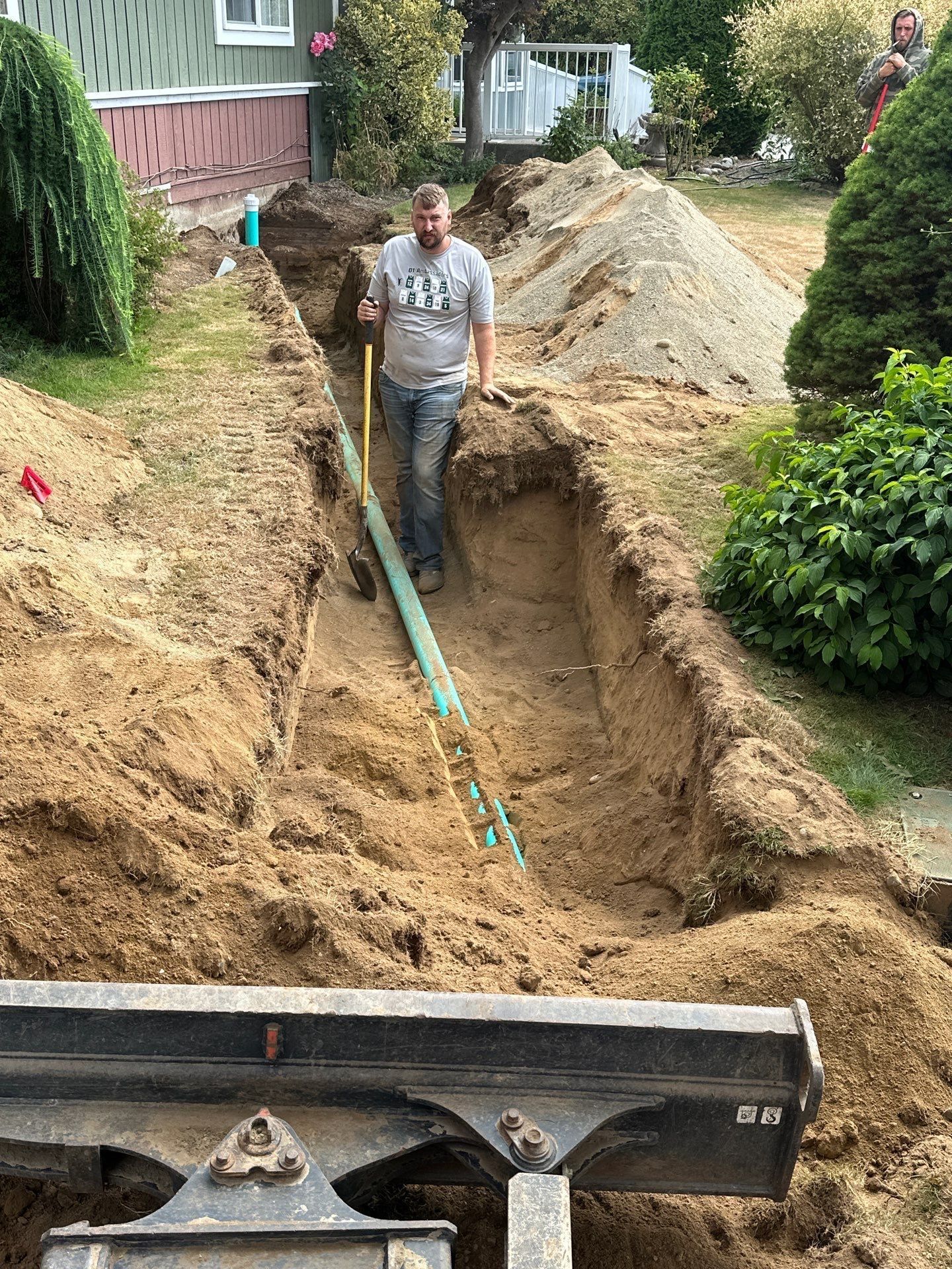 Worker in a trench beside a house, with excavation equipment and a large dirt pile nearby.