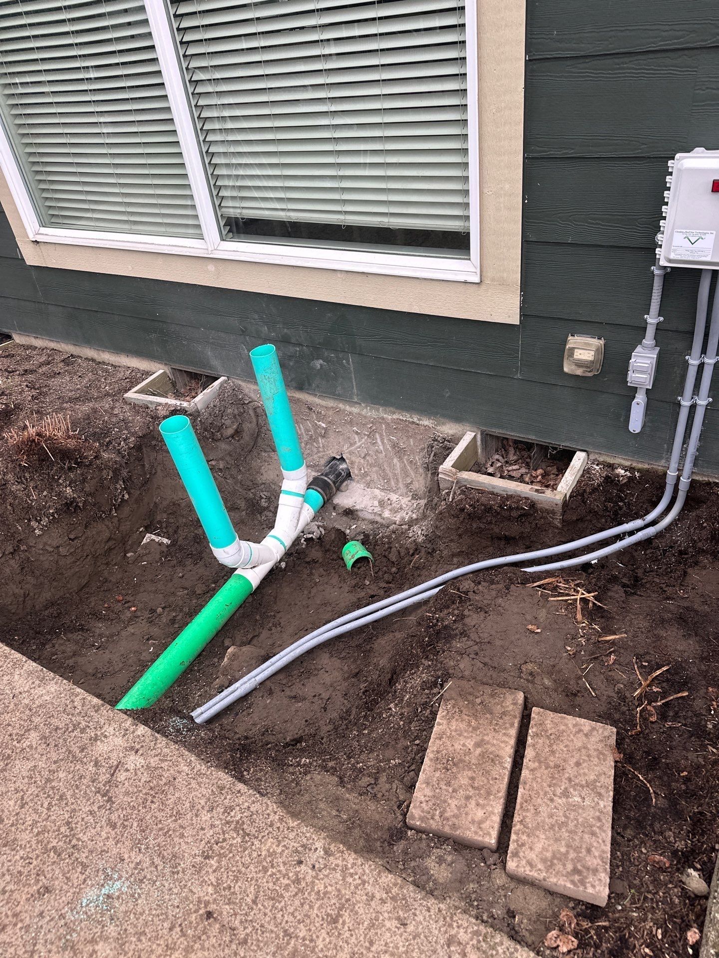 Outdoor utility pipes and conduits beside a house, with exposed green and gray pipes near a window.