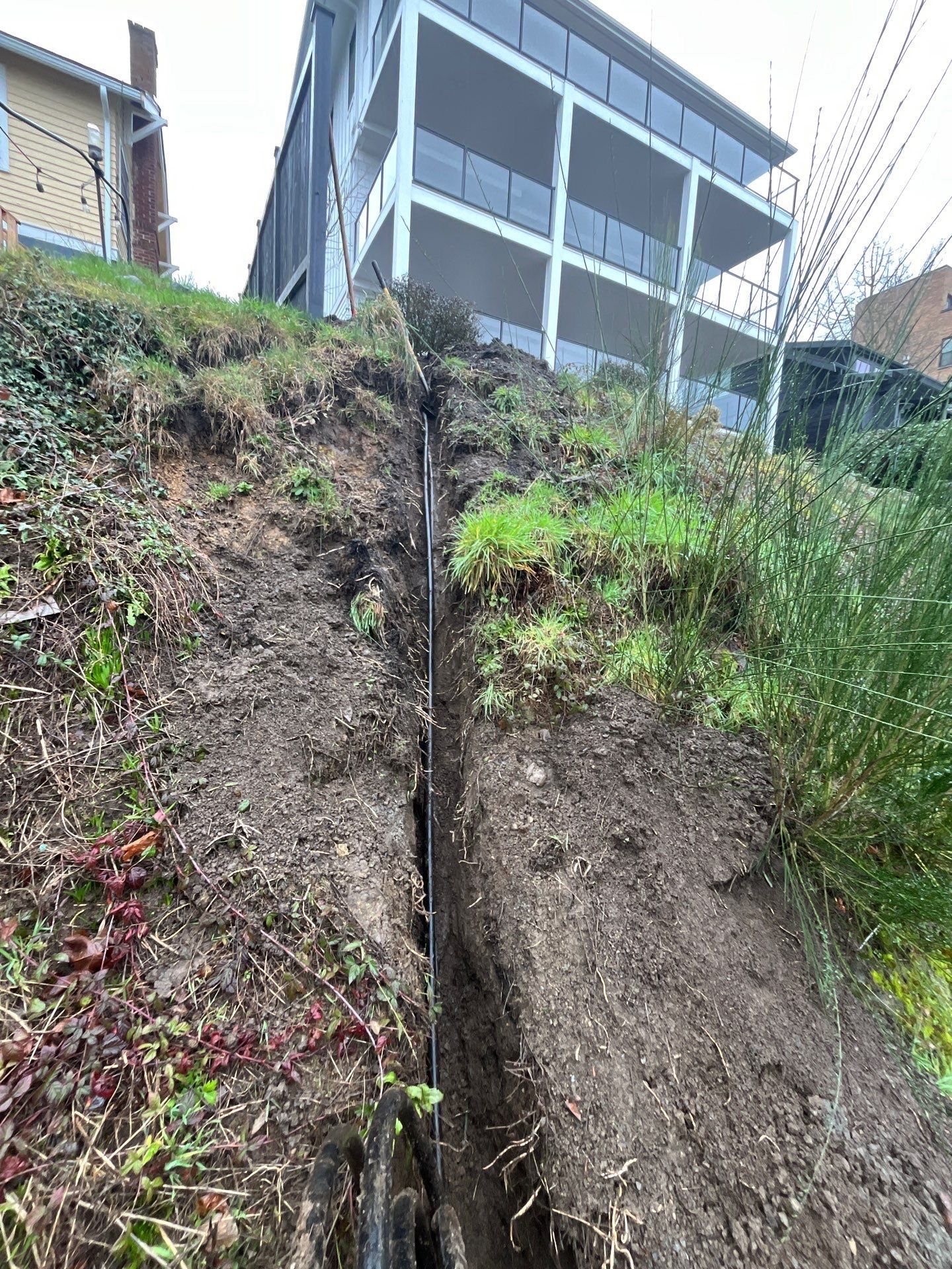 Steep dirt slope beside a building, with a narrow eroded trench and patchy green vegetation.