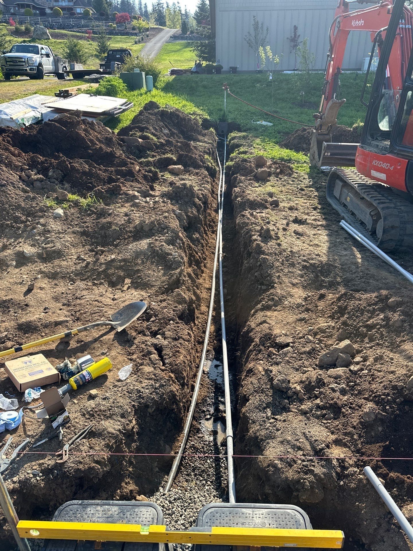 Excavator digging a long trench beside a house, with tools and dirt piled along the edges.