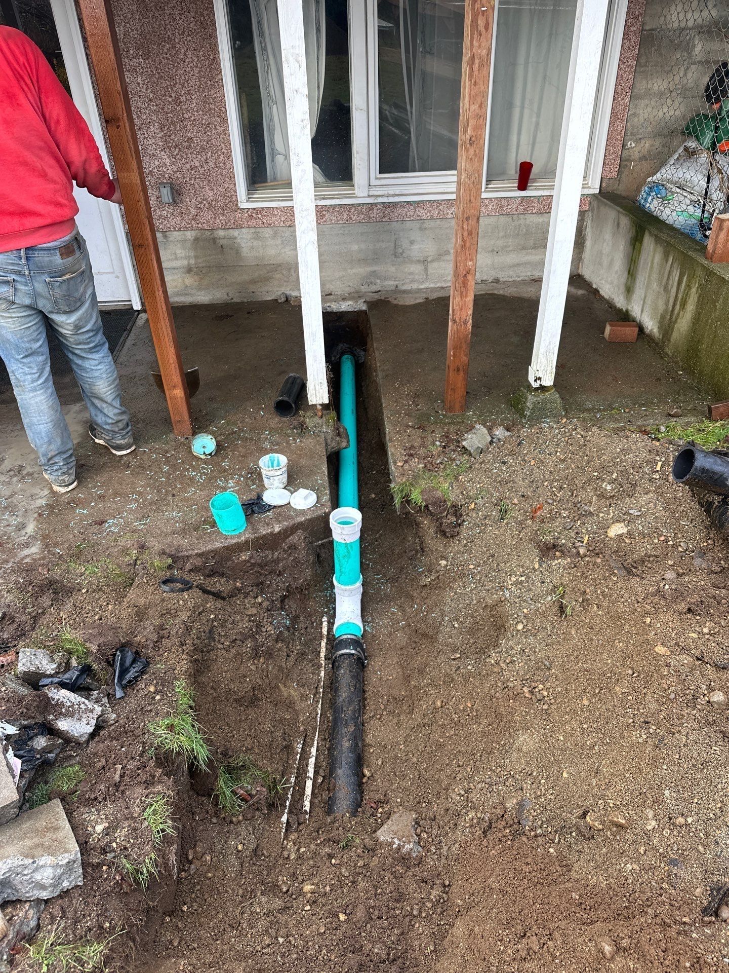 Exposed sewer pipes and fittings in a gravel yard outside a house window with two workers nearby.
