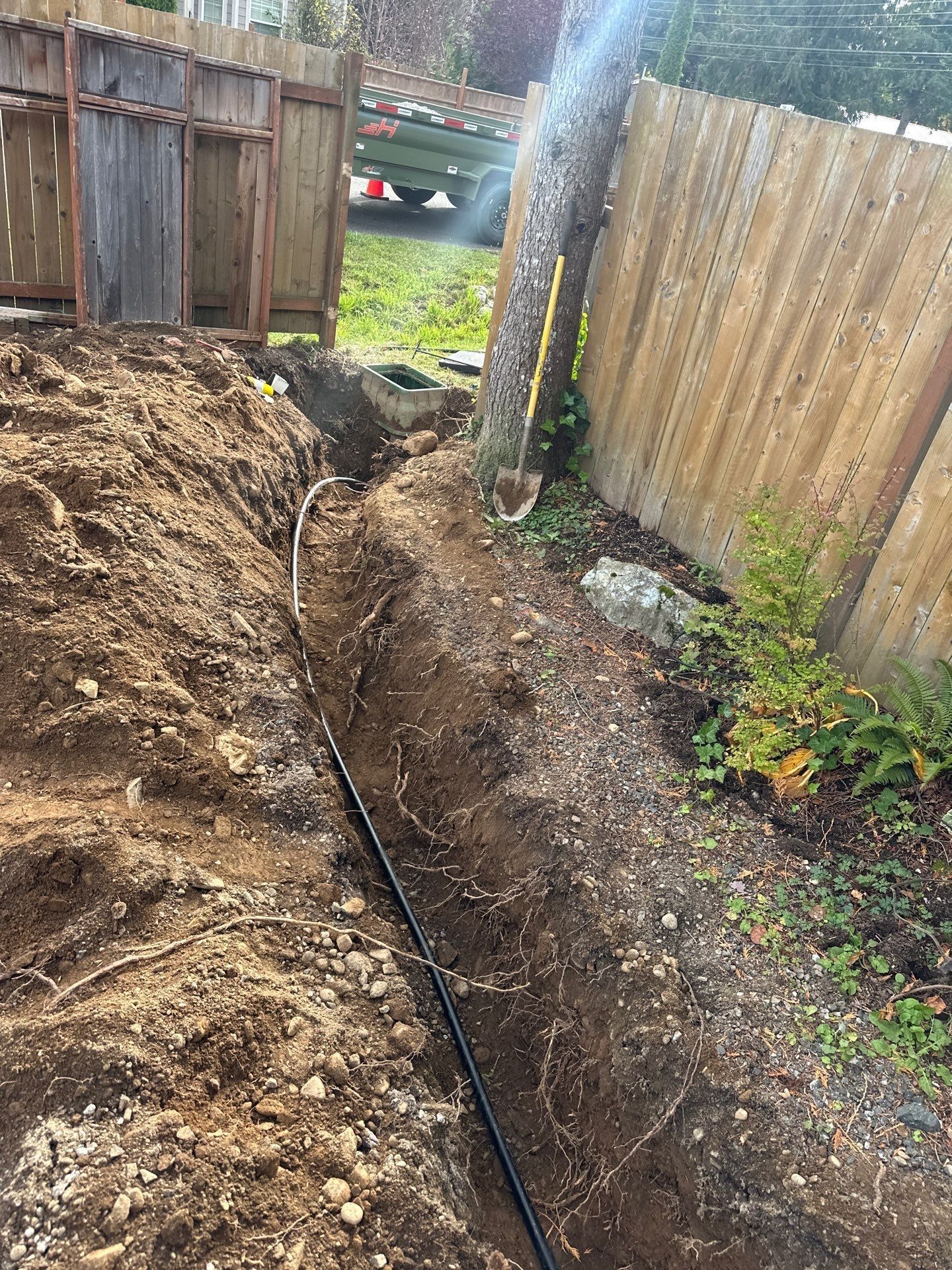 Narrow trench dug along a wooden fence, with exposed soil and a black cable running through it.