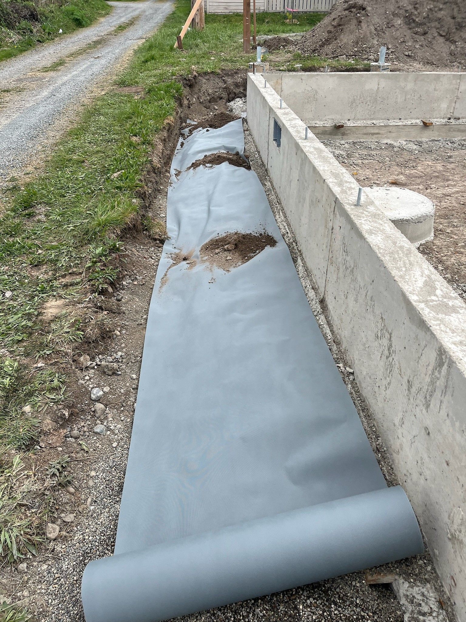 Blue landscape fabric lining a narrow trench beside a concrete foundation wall and gravel path.