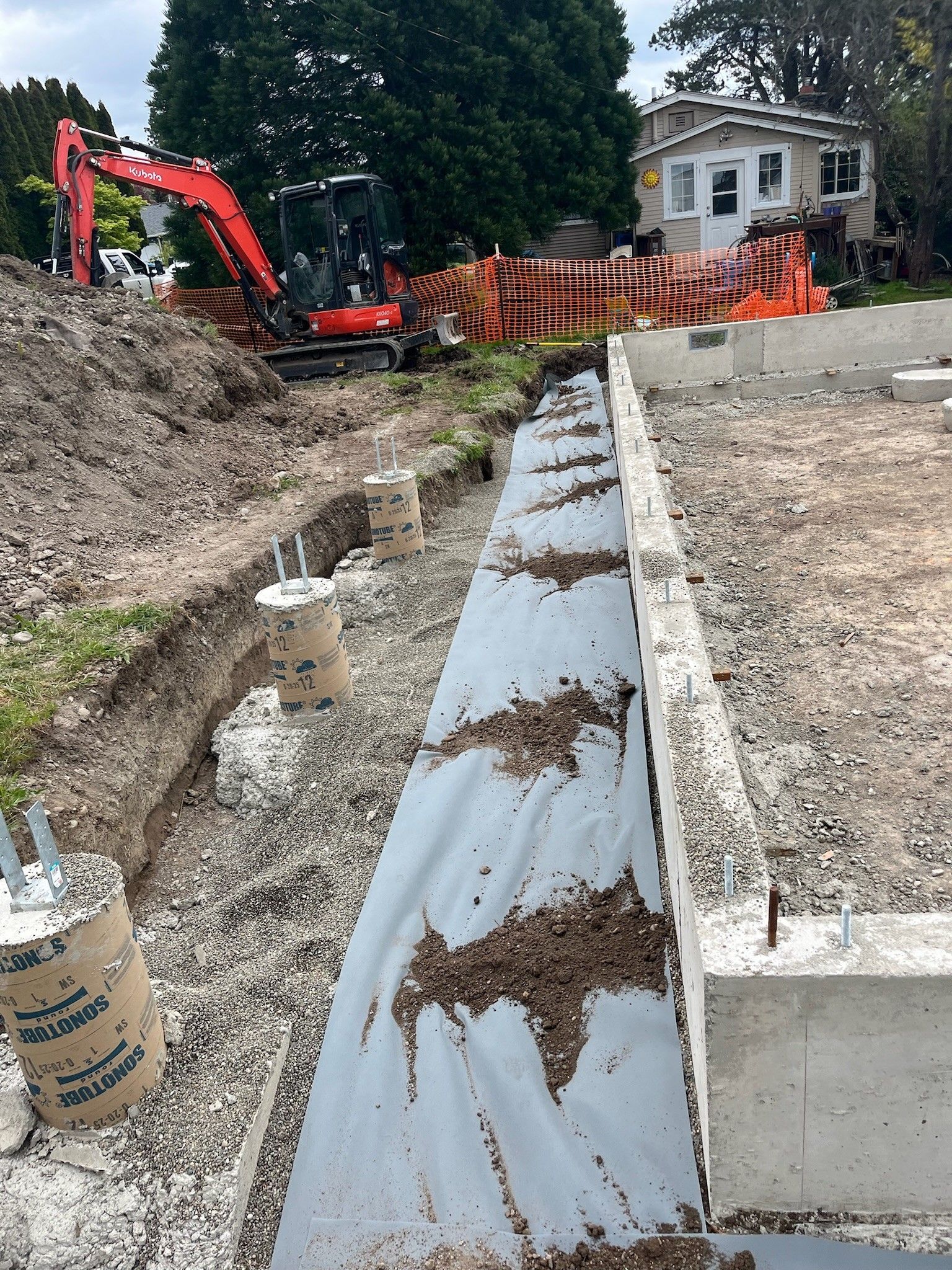 Excavator and orange safety fence at a construction site beside a narrow drainage trench and house.