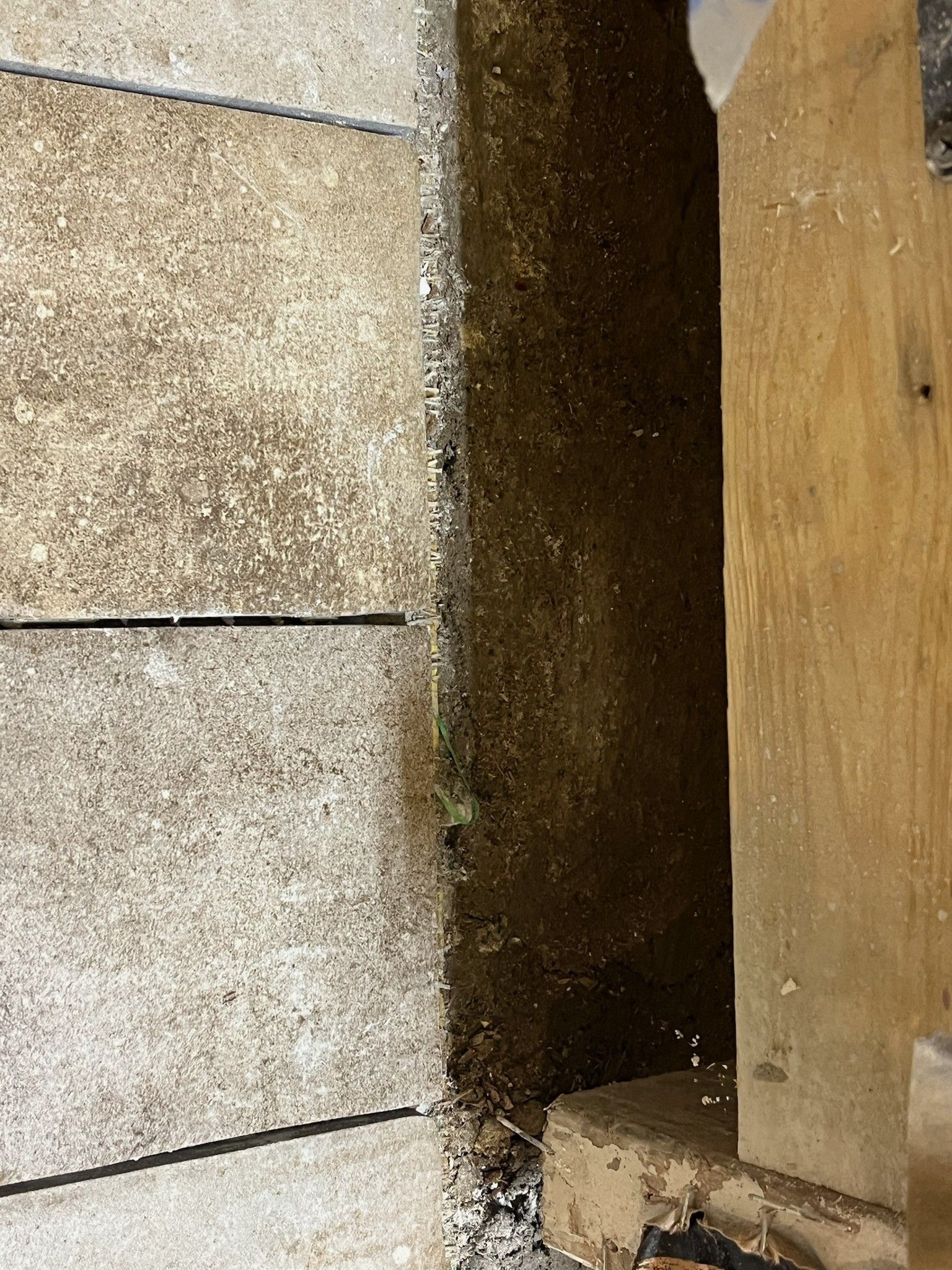 Narrow dark gap between a stone wall and a wooden panel, with debris at the base.