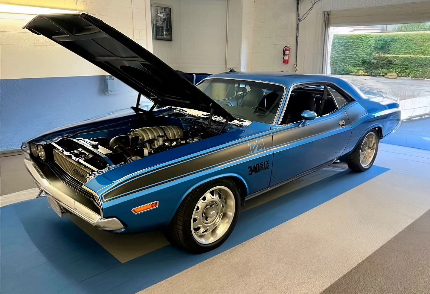 A blue dodge challenger with the hood up is parked in a garage.