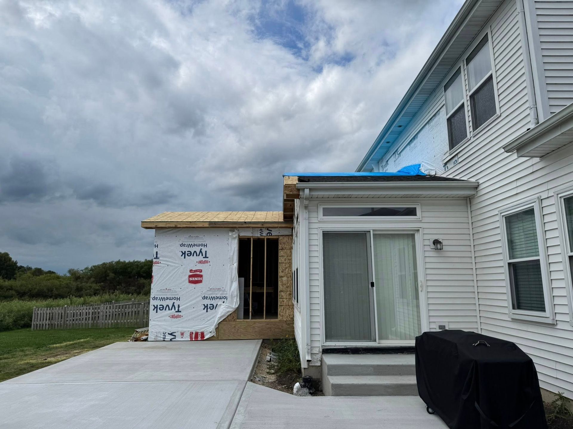 A house addition under construction. Gray sky, siding, framing, and a concrete patio are visible.
