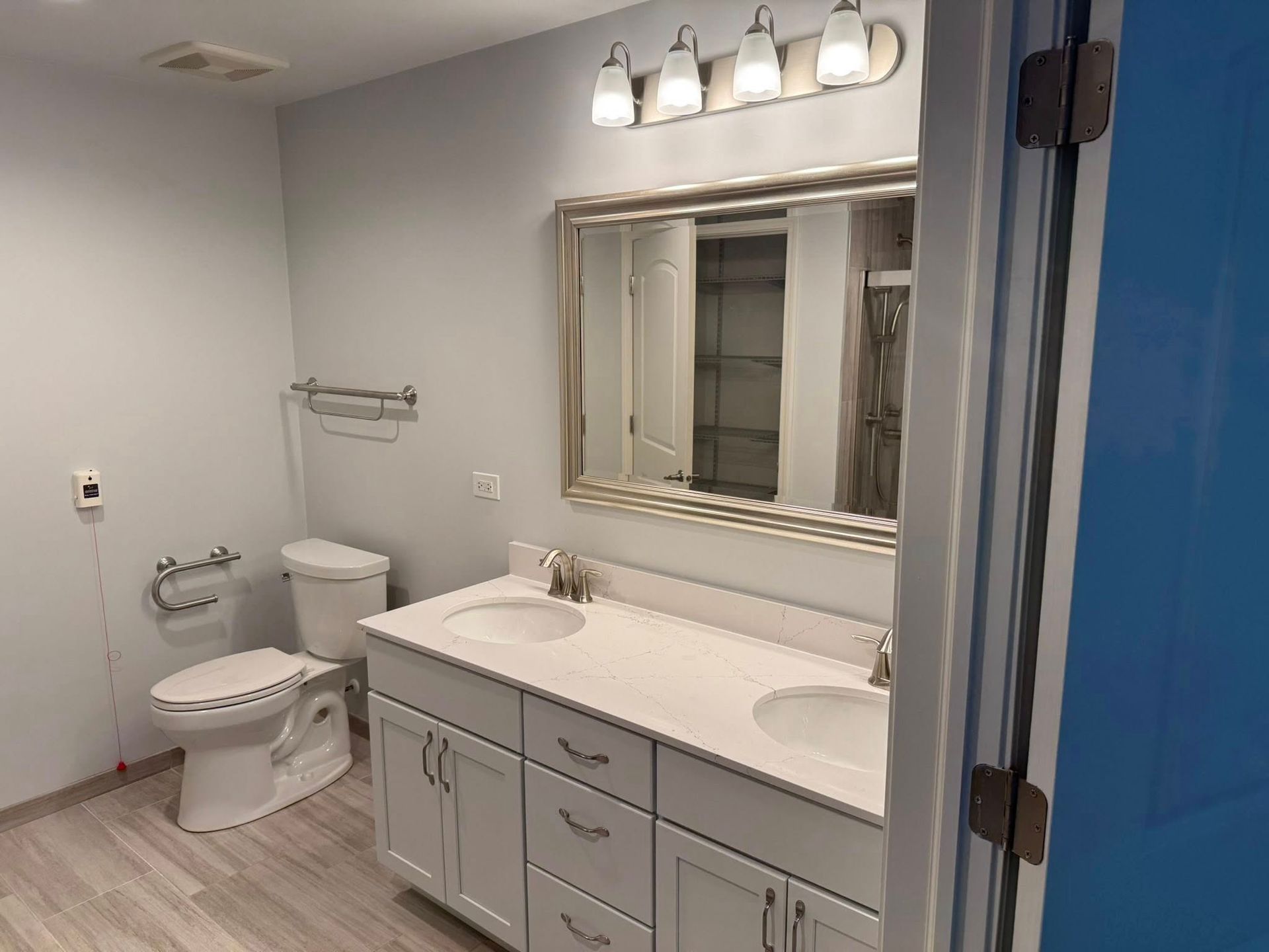 Bathroom with white double vanity, mirror, toilet, and silver fixtures. Light gray walls and wood-look floor.