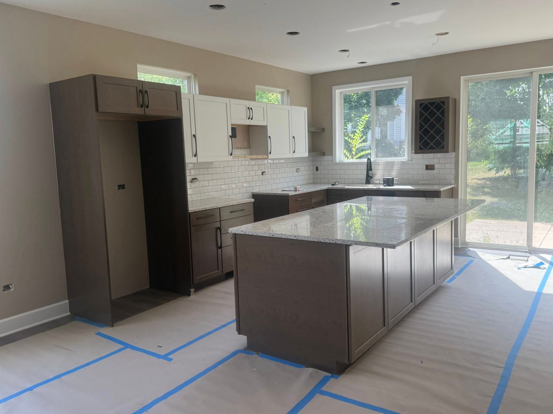 Kitchen under construction; dark brown cabinets, light countertops, island, light walls, blue tape on floor.