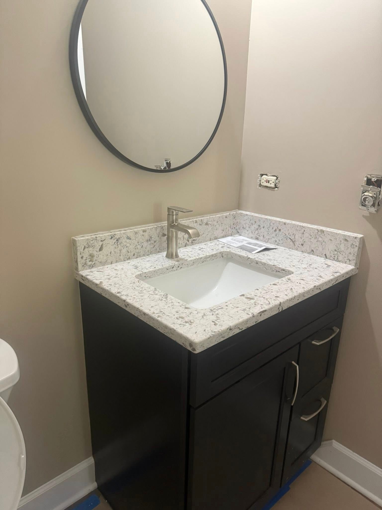 Bathroom vanity with black cabinet, speckled countertop, white sink, and round mirror.