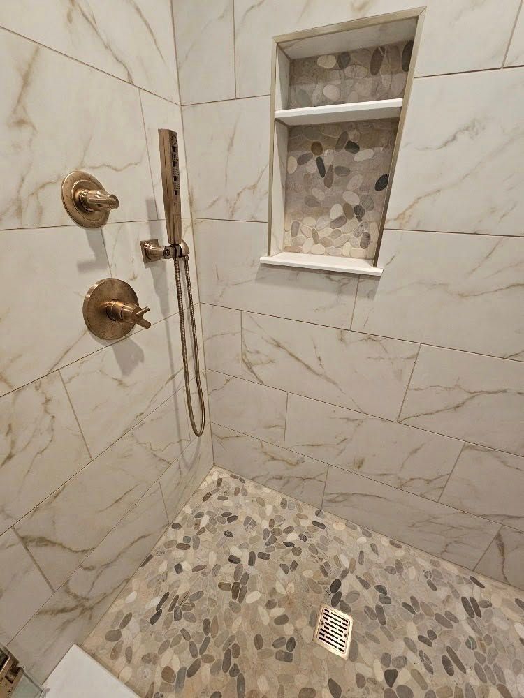 Shower with white marble tile walls, stone floor, and a built-in shelf with pebble-like tiles.