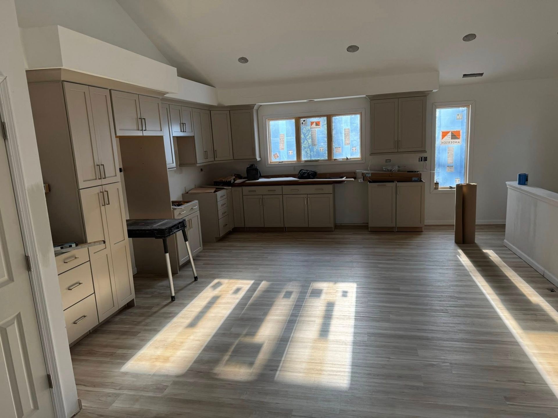 Bright empty kitchen with gray cabinets, large windows, and sunlight on wood floors.