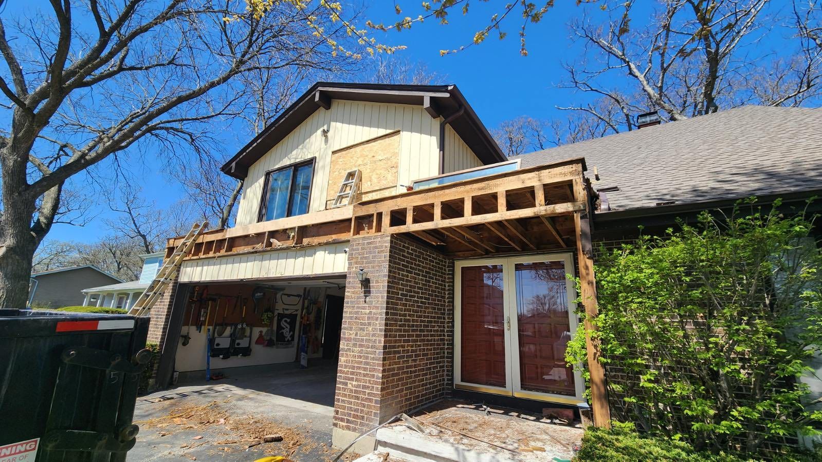 Stone-front suburban house with a covered porch, tan siding, and green shrubs under a clear blue sky