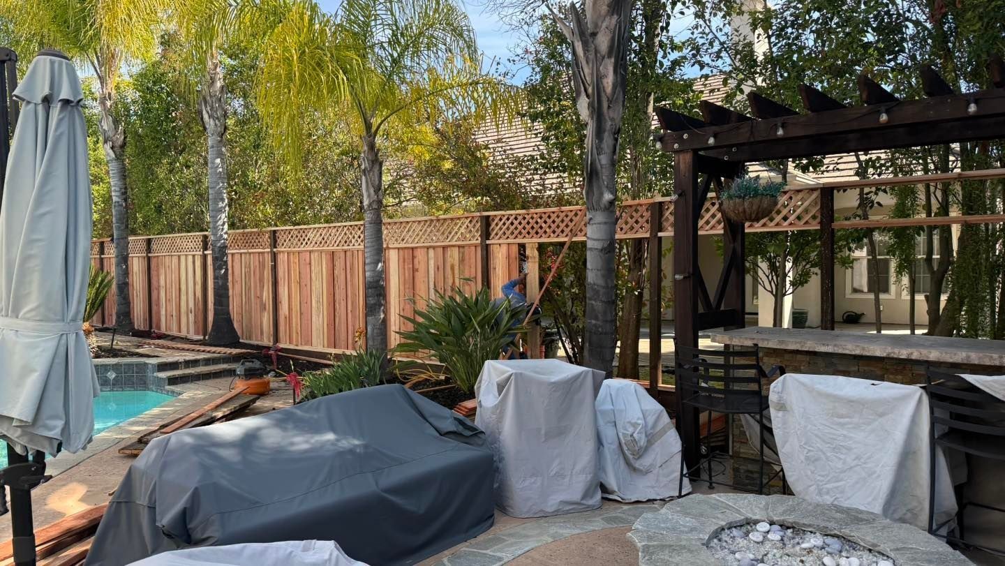 Backyard patio with covered furniture, pergola, pool, and wooden fence under trees