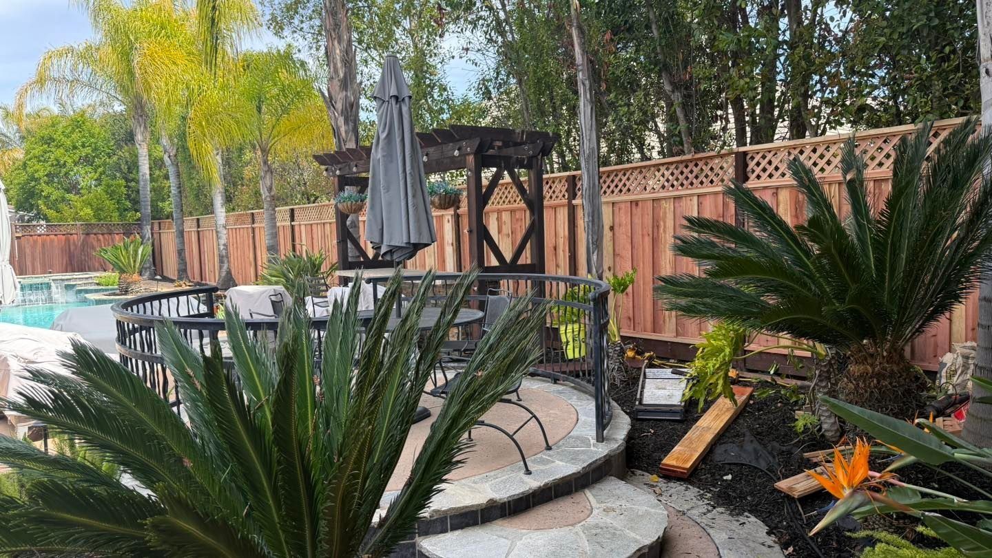 Patio with curved stone steps, outdoor seating, umbrella, and lush plants beside a wooden fence