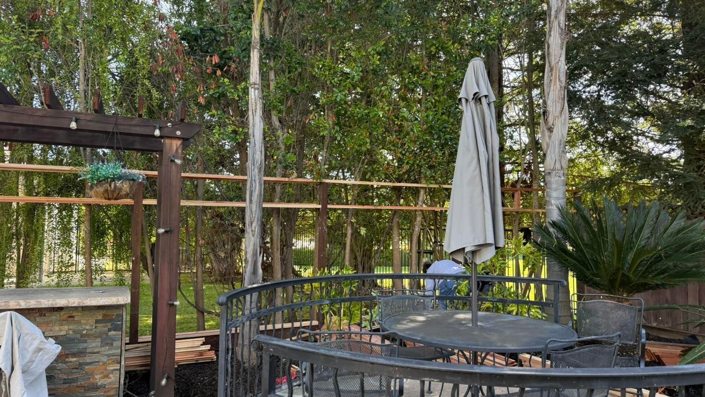 Outdoor patio with glass table, metal chairs, closed umbrella, and trees in the background.