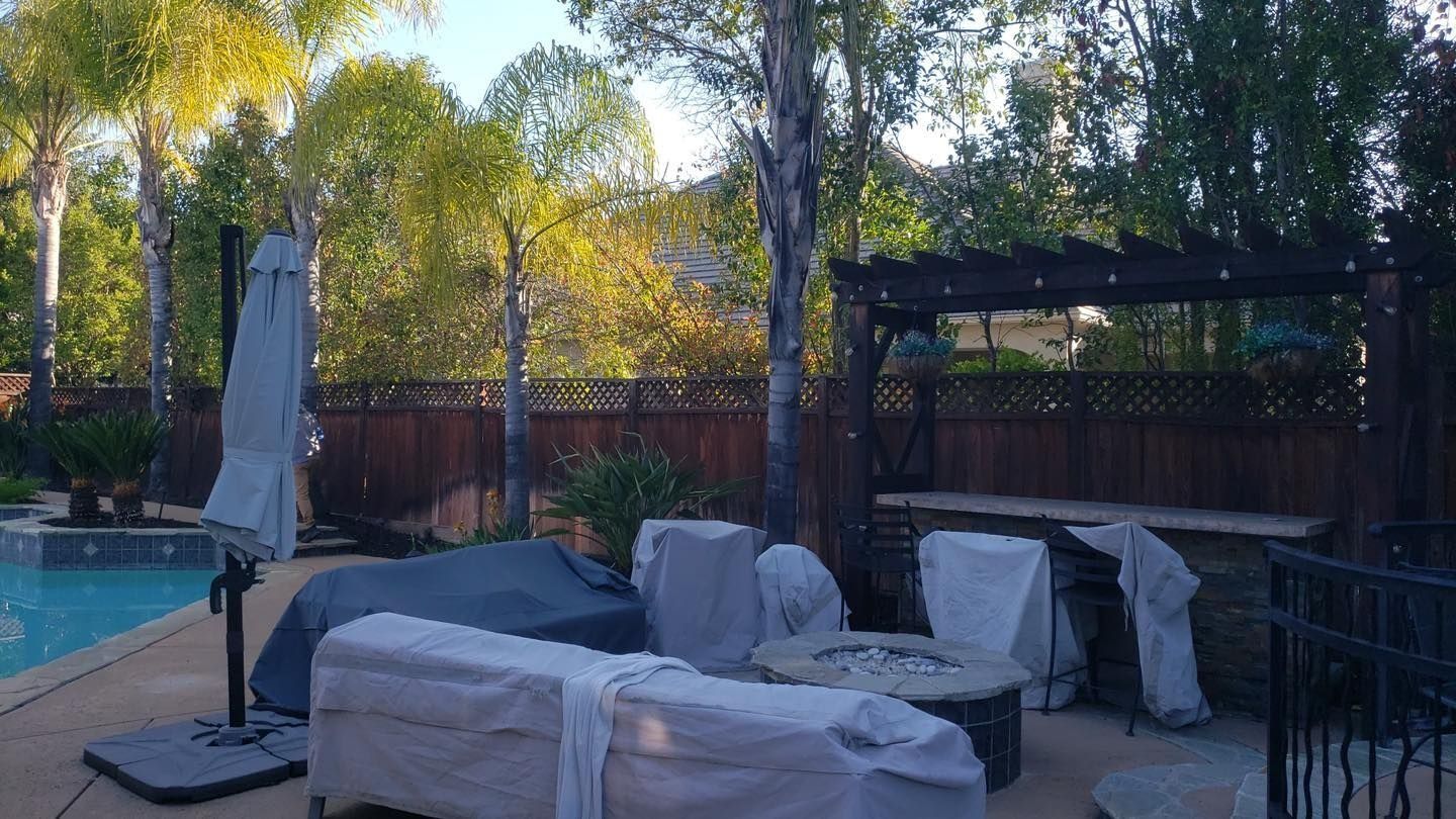 Backyard patio with covered outdoor furniture, pergola, and pool beside a wooden fence and trees.
