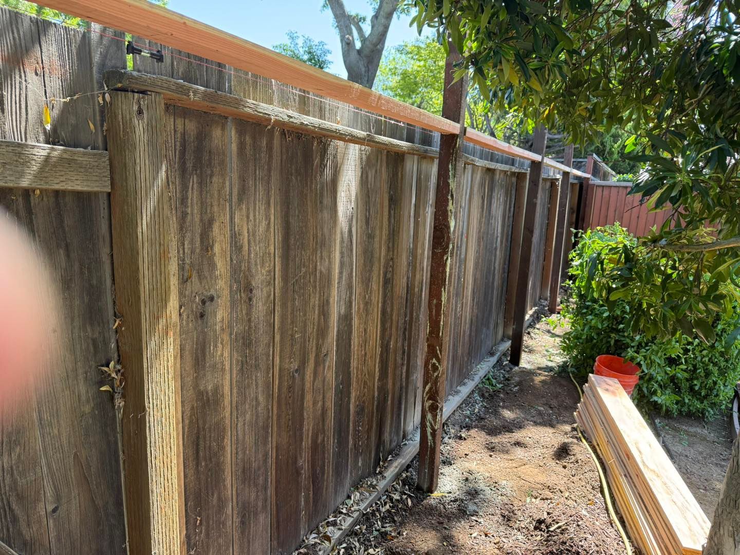 Weathered wooden fence beside a narrow dirt path, with greenery and a finger partially blocking the left side.