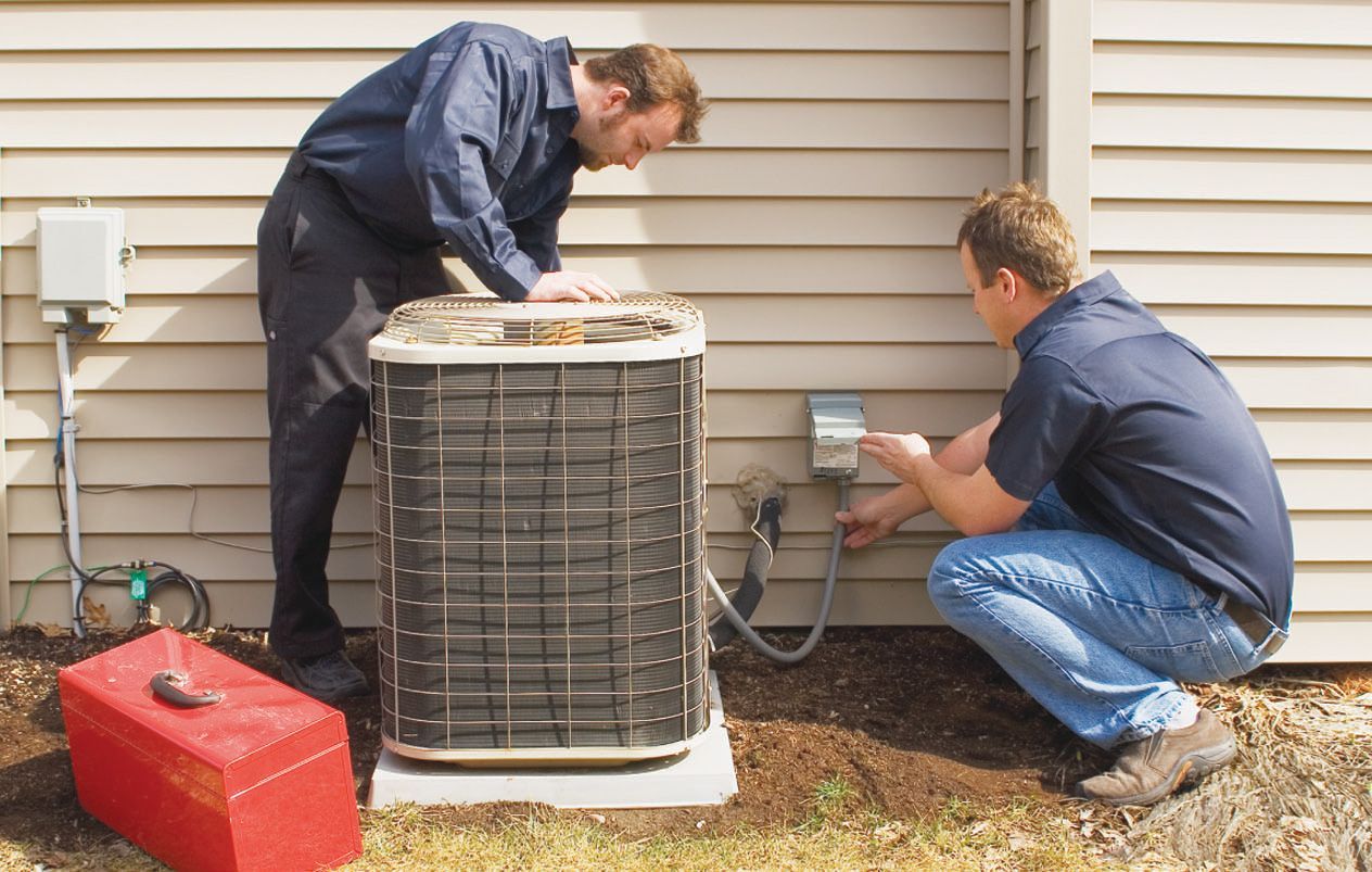 Two technicians in blue uniforms work on an air conditioner unit outside a building.