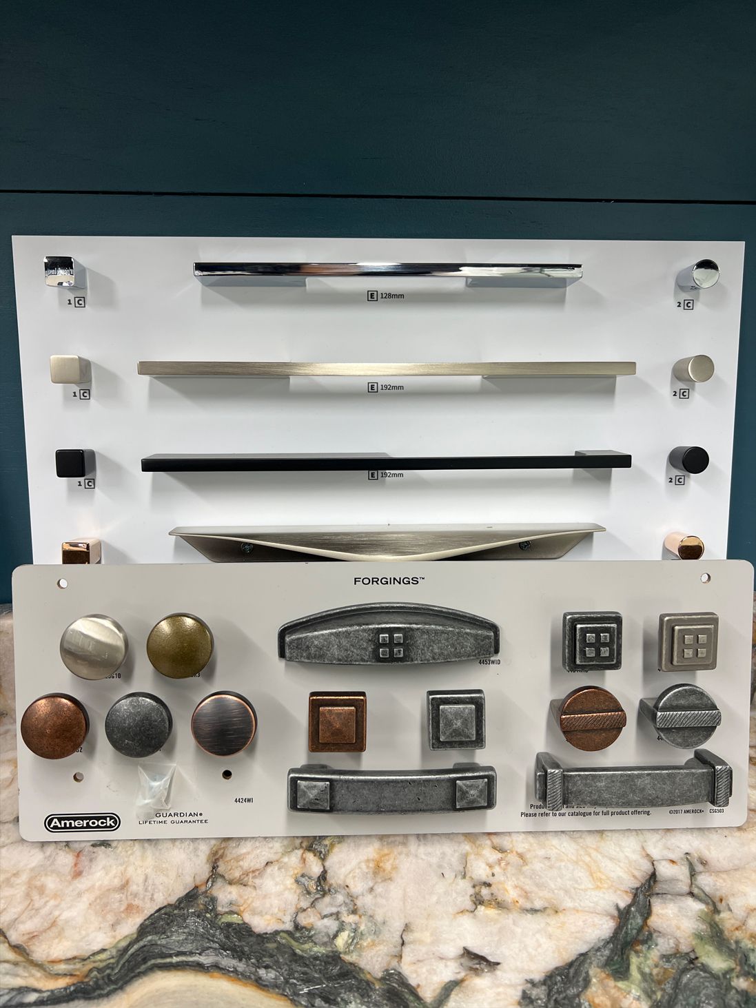 Displays of cabinet hardware in various styles and finishes on white boards against a turquoise wall.